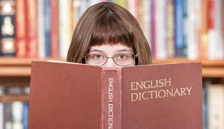 a girl with glasses peering over the top of an open English dictionary